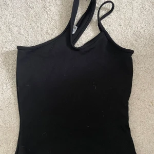 Svart asymmetrisk linnetopp från NA-KD - Säljer en svart linnetopp med asymmetriska axelband från NA-KD. Toppen är tight och har en stilren, minimalistisk look som passar perfekt till jeans eller kjol. Materialet känns mjukt och stretchigt, vilket gör den bekväm att bära hela dagen.