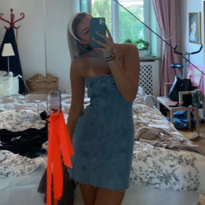 Blå axelbandslös jeansklänning - Säljer en snygg axelbandslös kort jeansklänning i blått. Klänningen har en rak passform och markerad midja, perfekt för dig som gillar en enkel men trendig look. Materialet är klassiskt denim och längden är ovanför knäna. Superfin till sneakers eller boots!