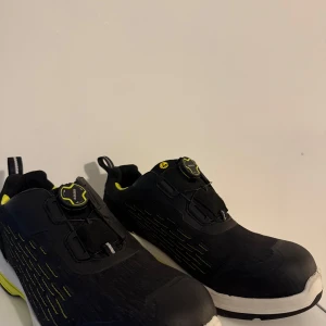 Svarta sneakers från Blåkläder med snabbsnörning - Svarta sneakers från Blåkläder med coola gula detaljer och vit sula. Skorna har snabbsnörning med BOA-system och sportig design. Insidan är fodrad i gult och yttermaterialet är syntet. Perfekta för dig som gillar stilrena och funktionella skor.