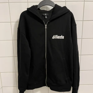 Svart hoodie med Atlanta-tryck H&M. Helt ny och oanvänd. - Svart hoodie från H&M med dragkedja och huva. Stor vit Atlanta-text på bröstet och ännu större tryck på ryggen. Relaxed fit för en chill vibe. Perfekt för dig som gillar streetstyle. Mjukt material och fickor framtill. Säljer på grund av att jag glömde att returnera varan. Passar inte riktigt min personliga stil längre. Köpte för 550 kr. Den är storlek S men skulle säga att den passar som M. 