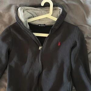 Svart hoodie från Polo Ralph Lauren - STRL= L (kids) passar någon som är ungefär 150-160 Svart hoodie från Polo Ralph Lauren med klassisk röd broderad logga på bröstet. Tröjan har dragkedja, huva med grå insida och långa ärmar. Tillverkad i mjukt bomull 