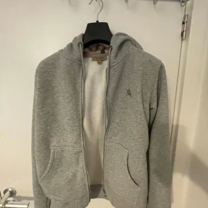 Grå hoodie från Burberry med huva - Säljer en grå hoodie från Burberry i 100% bomull. Tröjan har dragkedja, huva med klassiskt rutigt Burberry-foder och broderad logga på bröstet. Två stora fickor framtill och ribbade muddar. Perfekt för dig som gillar stilren och lyxig streetwear.