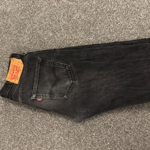 Svarta Levi's 501 jeans W30 L32 - Klassiska svarta Levi's 501 jeans med rak passform och femficksdesign. Jeansen har en snygg tvättad look och ikonisk läderpatch bak i midjan. Perfekta för dig som gillar tidlös stil och vill ha ett par jeans som funkar till allt.