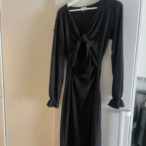 Svart långklänning med knyt framtill - Svart långklänning med långa ärmar och snygg knytning framtill som skapar en öppen detalj vid magen. Ärmarna har volang vid ärmslut och klänningen har en elegant, figurnära passform. Perfekt för dig som vill sticka ut med en cool och trendig look. Ett litet hål på ena ärmen.