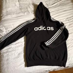 Svart Adidas hoodie med vita ränder - Svart hoodie från Adidas med klassiska vita ränder längs ärmarna och stor vit logga på bröstet. Tröjan har huva med snörning och en stor magficka. Perfekt för en sportig och avslappnad stil. Bra skick knappt använd.