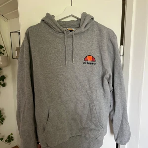 Grå hoodie från Ellesse med logga - Säljer en grå hoodie från Ellesse i storlek M. Tröjan har klassisk huva med snörning, magficka och Ellesse-logga broderad på bröstet. Perfekt för chill dagar och enkel att matcha med jeans eller joggers.