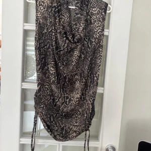 Kort blus med ormmönster Storlek L , 42-46 passar - Snygg kort blus med svart och beige ormmönster. Klänningen har dragsko i sidorna för justerbar längd och en lösare passform upptill. Perfekt för dig som vill sticka ut med ett djurmönster och coola detaljer.