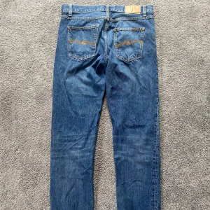 Blå jeans från Nudie Jeans Co 31/34 - Säljer ett par klassiska blå jeans från Nudie Jeans Co i storlek 31/34. Jeansen har raka ben, snygga kontrastsömmar och ikoniska bakfickor med broderad våg. Tillverkade i slitstarkt bomullsjeans, perfekta för en avslappnad och stilren look.