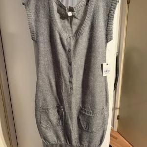 Grå stickad ärmlös klänning - Grå stickad klänning med v-ringning, knappar framtill och två stora fickor. Ärmlös modell med ribbade kanter och mjukt material som känns både chill och snygg. Perfekt att styla med boots eller sneakers för en cool look.