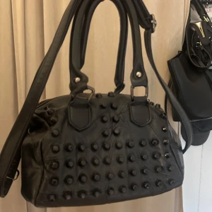 Svart handväska med nitar  -  svart handväska i skinn med massor av svarta nitar på framsidan. Väskan har både handtag och axelrem samt dragkedja upptill. Dragkedjan är ytterst lite missfärgad, men inget som märks i sin helhet. Dock finns det 1 innerficka med dragkedja, där det tillkommer ett hål i fickan. Men inget jag har haft problem med.💕