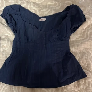 Mörkblå blus från Nelly med knappar - Supersöt mörkblå blus från Nelly med korta ärmar och v-ringning. Blusen har tre knappar framtill och en lätt figursydd passform som ger en feminin siluett. Perfekt att styla med jeans eller kjol för en trendig look.