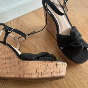 Svarta kilklackssandaler från H&M - Snygga svarta sandaler från H&M med hög korkliknande kilklack och bred knut framtill. Skorna har öppen tå, justerbar rem runt vristen med guldfärgat spänne och plattformssula. Perfekta för sommarens alla tillfällen när du vill ha lite extra höjd