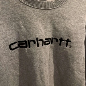 Grå Carhartt sweatshirt XS - Snygg grå sweatshirt från Carhartt i storlek XS. Tröjan har svart broderad logga framtill, rund halsringning och ribbade muddar vid ärmslut och nederkant. Perfekt basic att ha i garderoben för en clean och avslappnad stil. Säljer pga för liten.