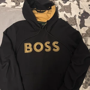 Svart hoodie från BOSS med guldtryck - Svart hoodie från BOSS i storlek M med stort guldigt BOSS-tryck på bröstet. Insidan av huvan är beige och har BOSS-logga längs kanten. Klassisk känguruficka och snörning i huvan. Perfekt för dig som gillar stilrena streetwear-plagg.