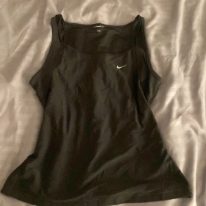 Svart linnetopp från Nike - Svart linnetopp från Nike med klassisk vit swoosh-logga på bröstet. Topp i sportig stil med breda axelband och rundad halsringning. Perfekt för träning eller chill dagar. Mjukt och stretchigt material som sitter skönt mot kroppen.