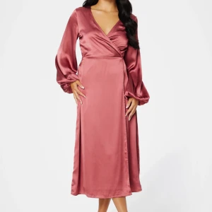 Rosa omlottklänning i satin - Snygg rosa långklänning i satin med omlottmodell och v-ringning. Klänningen har långa, puffiga ärmar och en elegant siluett som ger en lyxig känsla. Perfekt för dig som vill sticka ut med en feminin och trendig look. Helt oanvänd och ny, och den säljer jag för att jag råkade få med två stycken. Pris går att diskutera!