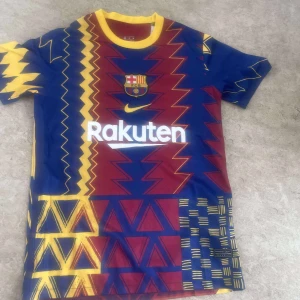 FC Barcelona x Nike mönstrad fotbollströja - Snygg FC Barcelona fotbollströja från Nike med unikt grafiskt mönster i blått, rött och gult. Tröjan har korta ärmar, gul krage och klubbmärke på bröstet. Rakuten-logga framtill. Tillverkad i lätt och ventilerande material, perfekt för match eller träning.