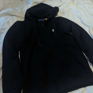Svart hoodie från Polo Ralph Lauren - Svart hoodie från Polo Ralph Lauren med dragkedja och huva. Klassisk broderad logga i vitt på bröstet och snörning i huvan. Mjuk bomullskänsla och långärmad modell, perfekt för chill dagar.