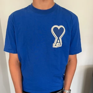 Blå t-shirt från Ami Paris med hjärtdetalj - Blå t-shirt från Ami Paris med stor vit hjärtdetalj och bokstaven A på bröstet. Klassisk rund hals och korta ärmar. Tillverkad i mjuk bomull, perfekt för en clean och stilren look. Tröjan är använd en gång och därför är skicket fof 10-10