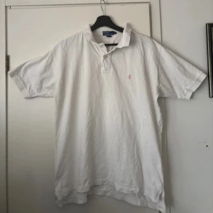 Vit pikétröja från Polo Ralph Lauren - Klassisk vit pikétröja från Polo Ralph Lauren med rosa broderad logga på bröstet. Tröjan har krage, knappar vid halsen och är tillverkad i 100% bomull. Perfekt för en clean och stilren look.