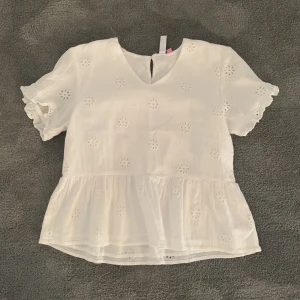 Vit blus med broderade blommor och volang.  - Söt vit blus med broderade blommor och volang nertill. Blusen har korta ärmar med spetsdetaljer och en liten v-ringning fram. Perfekt för varma dagar och ger en romantisk känsla till outfiten. Använd 1-2 gånger ! 
