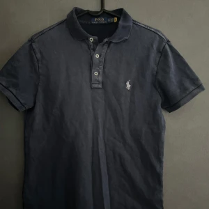 Mörkblå pikétröja från Polo Ralph Lauren - Säljer denna mörkblå pikétröja från Polo Ralph Lauren med vit broderad logga på bröstet. Tröjan har korta ärmar, krage och knappar framtill. Perfekt passform om du är mellan cirka 163-175. Du får med en snygg påse från polo vid köpet! Tröjan är helt oanvänd och väldigt snygg!