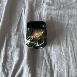 Svart AirPods-fodral med blixtmönster - Säljer ett svart AirPods-fodral med coolt blixtmönster i grönt, gult och rosa. Fodralet har en unik design som sticker ut och skyddar dina hörlurar på ett snyggt sätt. Perfekt för dig som vill ha något annorlunda till dina AirPods.