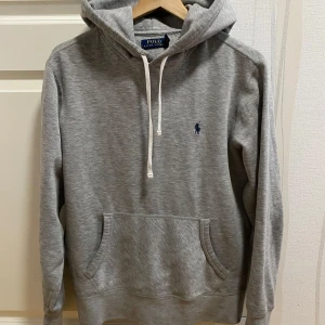 Polo Ralph Lauren Hoodie - Säljer en klassisk grå hoodie från Polo Ralph Lauren med broderad blå logga på bröstet. Tröjan har huva med vita snören och en stor magficka. Perfekt för en avslappnad stil.