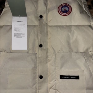 Beige dunväst från Canada Goose - Säljer en beige dunväst från Canada Goose med klassisk logotyp på bröstet och svarta knappar framtill. Västen har två stora fickor framtill och en diskret Canada Goose-label på ena fickan. Perfekt för lager-på-lager under kyliga dagar.