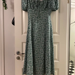 Grön blommig långklänning med puffärm - Säljer en grön långklänning med vitt blommönster och puffiga långa ärmar. Klänningen har knytning vid bysten och en slits framtill. Perfekt för vår och sommar, med en romantisk och trendig känsla.