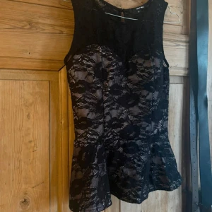 Svart spetsblus från Tally Weijl - Säljer en svart blus från Tally Weijl i storlek 38. Blusen har ett vackert spetslager med blommigt mönster och är figursydd med volang nertill. Den är ärmlös och har en rund halsringning samt knäppning bak. Ljusrosa innertyg. Jättefint skick och normal i storlek 