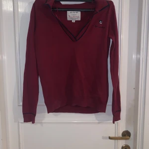 Vinröd långärmad tröja från Bondelid - Vinröd långärmad tröja från Bondelid med v-ringning och mörkblå kantdetaljer. Klassisk och stilren design med liten broderad logga på bröstet. Perfekt för dig som gillar en enkel men snygg look.
