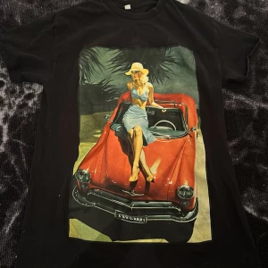 Svart t-shirt med retro bil- och pinup-tryck - Cool svart t-shirt med stort färgglatt tryck av en röd retrobil och en pinup-tjej på framsidan. Perfekt statement-plagg för dig som gillar vintage vibes och unika motiv. Då det är ett plagg från Revenite går det ej att köpa mer. Priset är inte hugget i sten.