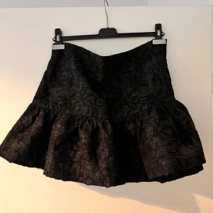 Svart brokad minikjol med volang - Säljer en svart minikjol i brokad från H&M. Kjolen har en volang nedtill som ger extra volym och struktur. Materialet har en lätt glansig yta med subtilt mönster. Perfekt för dig som vill ha en statementkjol med cool känsla.