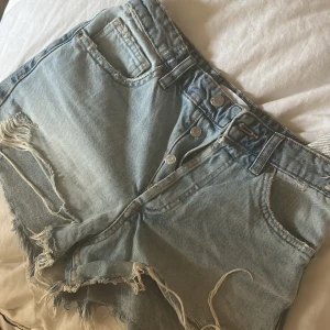 Ljusa jeansshorts med slitningar från ZARA - Säljer ett par ljusblå jeansshorts från ZARA med råa kanter och slitna detaljer både fram och bak. Klassisk femficksmodell med knappgylf och bälteshällor. Perfekta för varma dagar! Strl 34 i nyskick 