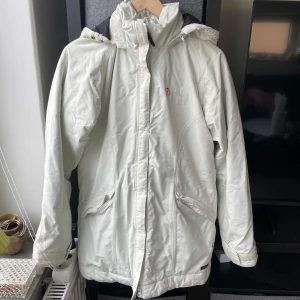 Vit vindjacka från Fjällräven - Säljer en vit och fodrad vindjacka från Fjällräven med huva och dragkedja framtill. Jackan har två fickor med dragkedja och broderad logga på bröstet. Perfekt för blåsiga dagar och enkel att matcha med olika outfits.