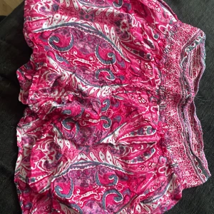 Mönstrade rosa shorts med smock - Färgglada shorts i rosa med vitt och blått paisleymönster. De har bred smock i midjan för en bekväm passform och är tillverkade i ett lätt bomullstyg. Perfekta för varma dagar när du vill ha något luftigt och snyggt.