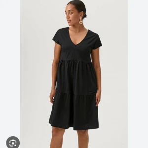 Svart v-ringad klänning med volang - En svart klänning med v-ringning och korta ärmar. Klänningen har en lös passform och volangdetaljer i kjolen som ger en luftig känsla. Perfekt för dig som gillar enkel och stilren design.