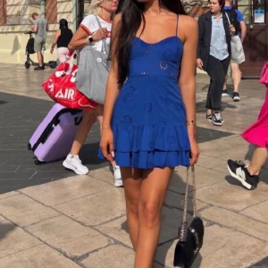 Blå kortklänning med volang - Snygg blå kortklänning med smala axelband och volangdetaljer nertill. Klänningen har broderade hålmönster och en feminin siluett. Perfekt för varma dagar och somriga tillfällen. Matcha med en liten väska för en komplett look.