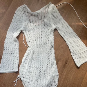 Vit stickad långärmad klänning med knyt - Säljer en vit stickad klänning med lång ärm och knyt i midjan. Klänningen har ett hålmönster och är figurnära med justerbar dragsko i sidan. Perfekt för sommarens varma dagar eller som strandplagg.