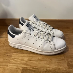Vita Adidas Stan Smith sneakers - Klassiska vita Adidas Stan Smith sneakers med mörkblåa detaljer på hälen och perforerade ränder på sidorna. Skorna har snörning och en platt sula, perfekt för en stilren och sportig look.