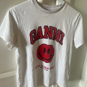 Vit t-shirt från Ganni med rött smileytryck - Vit t-shirt från Ganni med stort rött smileytryck och texten 'Have a nice day' framtill. Klassisk passform och rund halsringning. Perfekt för en avslappnad och cool stil.