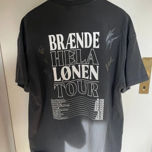 Svart t-shirt Brænde Hela Lønen Tour bolaget - Svart t-shirt med vit text på ryggen: 'BRÆNDE HELA LØNEN TOUR' och turnédatum. Framsidan har liten vit logga 'BOLAGET RECORDS' på bröstet. T-shirten har korta ärmar och flera autografer på ryggen. Klassisk passform och rund hals. Storlek M men är väldigt oversized. Passar som en L-XL