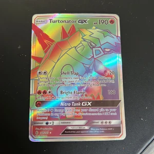 Turtonator GX Pokémonkort - Säljer ett holografiskt Turtonator GX-kort med 190 HP. Kortet har attacker som Shell Trap, Bright Flame och Nitro Tank GX. Framsidan är glansig med färgstark design och baksidan har klassiskt Pokémon-motiv.