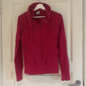 Rosa hoodie med dragkedja från Nike - Säljer en rosa hoodie från Nike med dragkedja framtill och huva med snörning. Jackan har långa ärmar och två fickor med dragkedja. Perfekt för träning eller chill dagar.