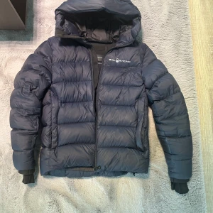 Mörkblå dunjacka från Sail Racing - Säljer en mörkblå dunjacka från Sail Racing med huva och dragkedja. Jackan har en puffig design och logga på bröstet. riktigt skön att bära. Inga skador eller avvikelser! Kvitto finns! Original pris 4000kr 