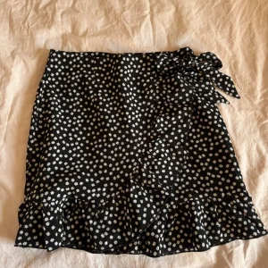 Svart omlottkjol med vita blommor - Supersöt svart omlottkjol med små vita blommor och volangkant. Kjolen har en superfin knytning i midjan!!