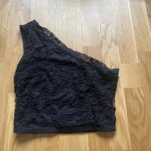 Svart spets one shoulder topp - Säljer en svart one shoulder topp i spets med blommigt mönster. Perfekt för dig som vill ha en trendig och elegant look. Toppen har en asymmetrisk axel och är figurnära.