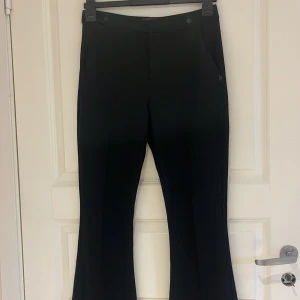 Svarta kostymbyxor från Scotch & Soda - Stilrena svarta kostymbyxor från Scotch & Soda med bootcut-passform. Byxorna har diskreta fickor bak och klassisk design, perfekta för en dressad look. Materialet känns mjukt och följsamt. Lite glittrigt tyg