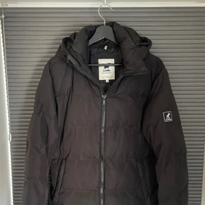 Svart dunjacka från Fat Moose - Säljer en svart dunjacka från Fat Moose med huva och dragkedja framtill. Jackan har en stilren design med diskret logga på ärmen och är perfekt för kalla dagar. Passformen är normal och jackan är vadderad för extra värme.